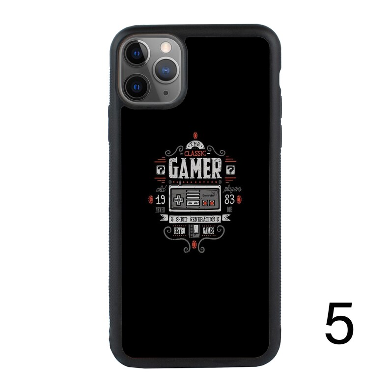Puede incluir: Carcasa de tel&eacute;fono negra con un dise&ntilde;o de juego retro. El dise&ntilde;o presenta un mando de videojuego vintage con el texto "The Classic Gamer" y "8-Bit Generation of Retro Games".