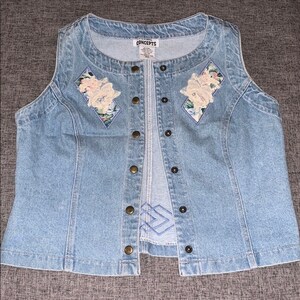 May include: A light blue denim waistcoat with a zip fastening and button accents. The waistcoat features floral embroidery on the front with a diamond-shaped design. The waistcoat has the text "Concepts" on the label.