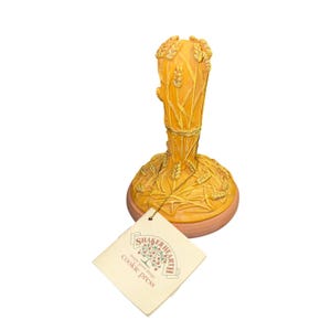 May include: A yellow ceramic cookie press featuring a raised wheat stalk design on the handle and base. A square paper tag is attached, printed with the text Shaker Hearth simply good things cookie press and a tree logo in a rectangular frame.
