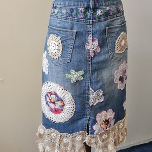 May include: A denim skirt with a back zip and a row of embroidered flowers along the waistband. The skirt is decorated with various crocheted lace appliques and a ruffled lace trim at the hem.