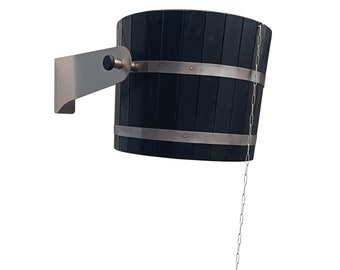 Può includere: Un secchio di legno nero con fasce metalliche e una catena che pende da esso. Il secchio è attaccato a un braccio metallico che è montato su un muro.