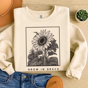 Op de afbeelding: Crèmekleurige sweatshirt met een zwart-witte zonnebloemillustratie in een zwarte rechthoek. De tekst "GROW IN GRACE" staat eronder. De sweatshirt wordt gecombineerd met een blauwe jeans en een bruine sandaal.
