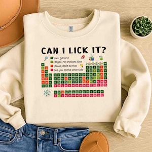 May include: Cream-colored sweatshirt with the text "CAN I LICK IT?" and a periodic table design. The design includes a color-coded key indicating different levels of caution. A fun and educational science-themed apparel item.