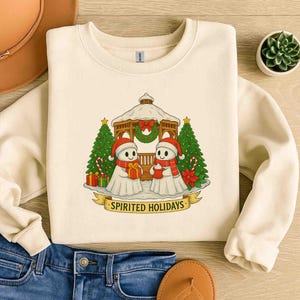 May include: A cream-coloured sweatshirt with a festive graphic. The design shows two snowman-like figures in Santa hats and scarves, in front of a gazebo and Christmas trees. The text "SPIRITED HOLIDAYS" is on a banner below the figures.