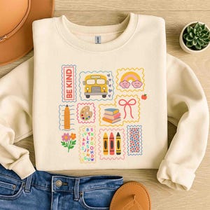 Puede incluir: Sudadera de color crema con gráficos coloridos con temática escolar. Presenta un autobús escolar amarillo, un arcoíris, crayones y las palabras "BE KIND". El diseño está impreso en una tela suave y cómoda.
