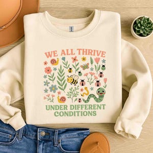 May include: Cream-colored sweatshirt with a colorful graphic of insects, flowers, and the text "WE ALL THRIVE UNDER DIFFERENT CONDITIONS" in pink and green. The design includes bees, ladybugs, snails, and a caterpillar.