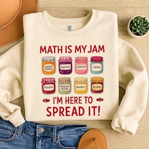 Può includere: Felpa color crema con la scritta "MATH IS MY JAM" e "I'M HERE TO SPREAD IT!". Il design presenta barattoli etichettati con concetti matematici come Frazioni, Geometria e Decimali. La felpa è abbinata a jeans blu e un sandalo.