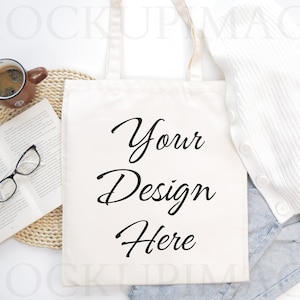 May include: A white canvas tote bag with the text "Your Design Here" in black cursive writing. The bag is lying on a white surface with a book, a cup of coffee, and a white sweater.