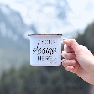 Könnte beinhalten: Weiße Emaille-Campingtasse mit schwarzem Henkel und dem Text "YOUR design HERE" in schwarzer Schrift. Die Tasse wird von einer Hand vor einem verschwommenen Hintergrund aus Bergen gehalten.