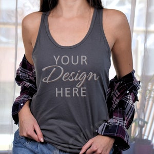 Puede incluir: Una camiseta sin mangas gris oscuro con el texto "YOUR Design HERE" en una fuente cursiva. Una camisa a cuadros en tonos burdeos, negro y blanco está drapeada sobre los hombros. La modelo lleva vaqueros azules.