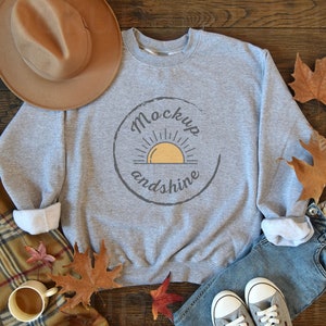 Pode incluir: Camisola cinzenta com um gráfico de um sol e o texto "Mockup and shine" num círculo. A camisola está colocada numa superfície de madeira com um chapéu, calças de ganga e folhas de outono.