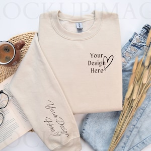 May include: A beige sweatshirt with the text "Your Design Here" and a heart on the front. The sweatshirt is lying on a white surface with a book, a cup of coffee, a pair of glasses, and a pair of jeans with dried grass.