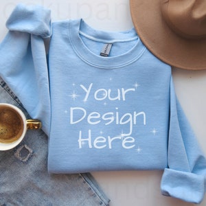 May include: A light blue sweatshirt with the text "Your Design Here" in white. A brown hat and a cup of coffee with a gold handle are also visible. The sweatshirt is folded on top of a pair of blue jeans.