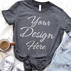 May include: A dark gray t-shirt with the text "Your Design Here" in white. The t-shirt is laying on a white surface with a pair of light blue jeans, a white sweater, and a brown coffee mug.