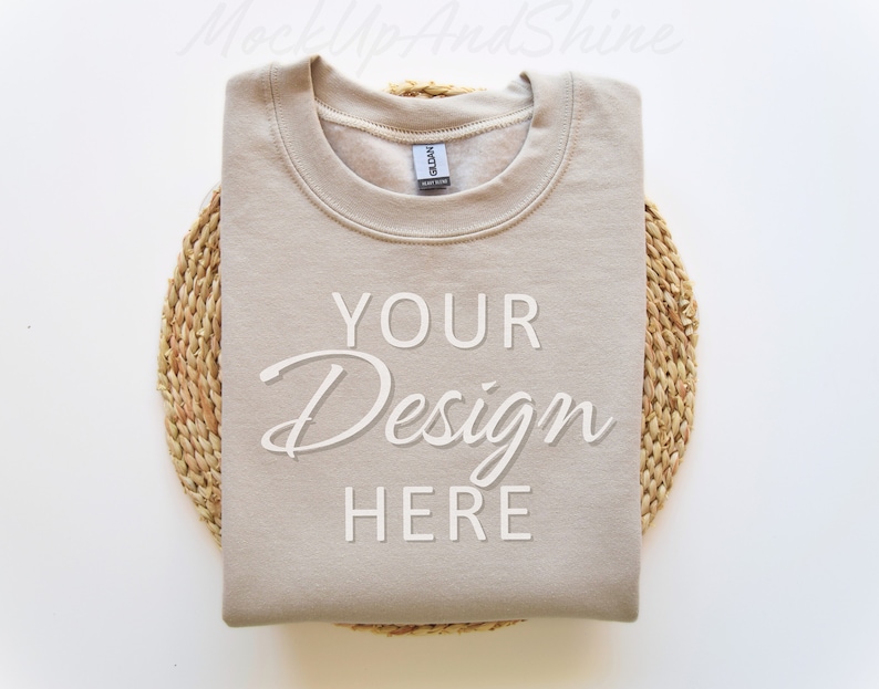 Peut inclure: Un sweat-shirt beige avec le texte "YOUR Design HERE" imprim&eacute; sur le devant. Le sweat-shirt est pli&eacute; et plac&eacute; sur un tapis en paille tress&eacute;e.