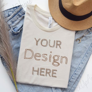 May include: A beige t-shirt with a ribbed crew neck and a brown outline of the text "YOUR Design HERE" on the front. The t-shirt is laid out on a white surface with blue jeans and a straw hat.