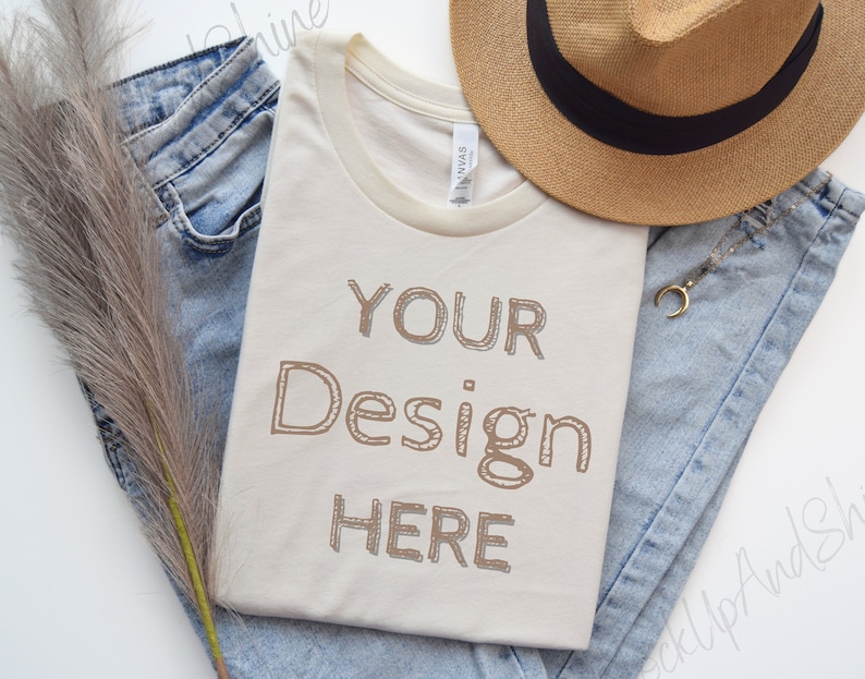 May include: A beige t-shirt with a ribbed crew neck and a brown outline of the text "YOUR Design HERE" on the front. The t-shirt is laid out on a white surface with blue jeans and a straw hat.