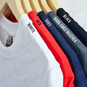 Puede incluir: Cinco camisetas colgadas en perchas de madera. Las camisetas son de los siguientes colores: blanco, rojo, azul marino, gris oscuro jaspeado y negro. El color de cada camiseta está escrito en la etiqueta.