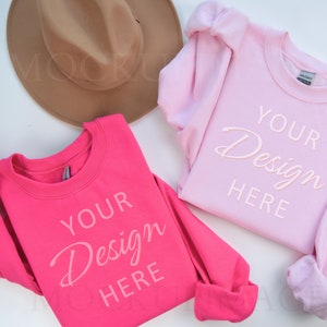 Könnte beinhalten: Zwei Sweatshirts, eines rosa und eines pinkfarben, liegen flach auf einer weißen Oberfläche. Beide Sweatshirts haben den Text "Your Design Here" auf der Vorderseite aufgedruckt. Ein brauner Fedora-Hut ist ebenfalls auf dem Bild zu sehen.