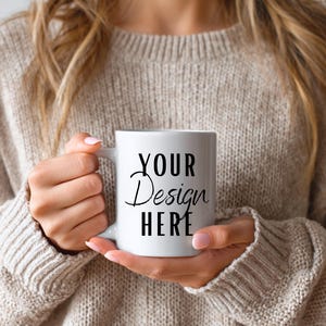 Puede incluir: Una taza de cerámica blanca con el texto "YOUR Design HERE" en escritura negra y letras de imprenta. La taza está sostenida por una persona que lleva un suéter beige.