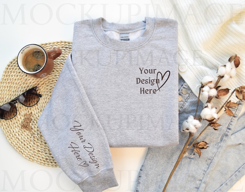 May include: A gray sweatshirt with the text "Your Design Here" on the front and "Your Design Here" on the left sleeve. The sweatshirt is laid flat on a white surface with a pair of jeans, a white cardigan, and a cotton branch.