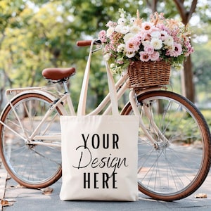 May include: A cream-colored tote bag with the text "YOUR Design HERE" is propped against a vintage bicycle. The bicycle has a brown leather seat and a wicker basket filled with pink and white flowers. The scene is set outdoors.