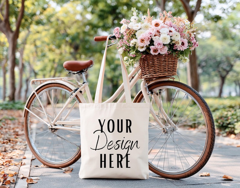 May include: A cream-colored tote bag with the text "YOUR Design HERE" is propped against a vintage bicycle. The bicycle has a brown leather seat and a wicker basket filled with pink and white flowers. The scene is set outdoors.