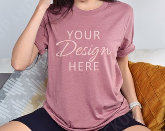 Può includere: Una donna indossa una maglietta color malva con la scritta "YOUR Design HERE" stampata sul davanti. È seduta su un divano con una coperta e un cuscino.