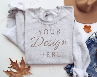 May include: A gray sweatshirt with the text "YOUR Design HERE" on the front. The sweatshirt is laid flat on a white surface with a brown hat, blue jeans, and fall leaves.