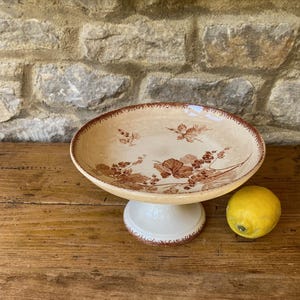 May include: A vintage cake stand in cream with a brown floral pattern. It features a wide, shallow dish and a short, pedestal base. A yellow lemon rests beside the stand on a wooden surface.