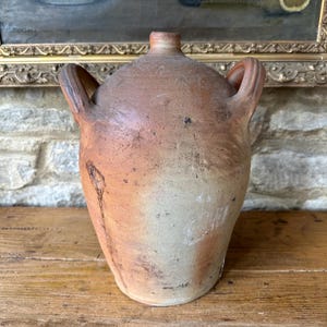 May include: A rustic, antique brown stoneware jug with two side handles. The jug has a rounded body and a narrow neck. The surface shows variations in color, with lighter and darker areas. The jug is sitting on a wooden surface.