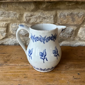 Puede incluir: Jarra de cerámica blanca con diseños florales azules. La jarra tiene un asa curva y un pico. El patrón floral azul decora el borde y el cuerpo. La jarra está sobre una superficie de madera.
