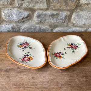 Könnte beinhalten: Zwei cremefarbene, unregelmäßig geformte Keramikplatten mit Blumendesigns. Jede Platte zeigt ein gemaltes Blumenarrangement in Rot-, Orange-, Lila- und Grüntönen. Die Ränder sind mit einem orangefarbenen Rand versehen. Die Teller werden auf einer Holzoberfläche präsentiert.
