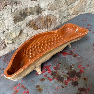May include: A long, narrow, fish-shaped ceramic mold with a glossy, deep orange-brown glazed interior featuring embossed scale patterns. The exterior is a matte, unglazed beige color, and the vessel is supported by three small, short feet.