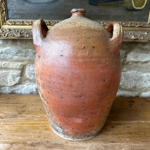 May include: A rustic, antique stoneware jug with a reddish-brown hue and two arched handles. The jug has a rounded body and a narrow neck with a small spout. The surface shows signs of age and wear, adding to its vintage charm. The jug is likely used for storing liquids.