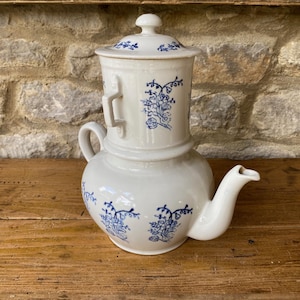 Op de afbeelding: Witte keramische koffiepot met blauwe bloemmotieven. De pot heeft een cilindrische bovenkant met deksel, een afgeronde basis, een handvat en een tuit. Staat op een houten oppervlak.