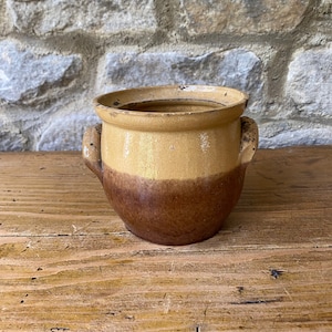May include: An antique ceramic pot with a rounded body and two small handles. The upper section is a light tan, fading into a rich brown at the base. The pot has a rustic, aged appearance, suggesting it is vintage and has a unique charm.