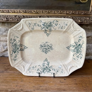 May include: Cream-colored rectangular serving platter with a floral design in teal. The platter has a slightly raised edge and is supported by a black metal stand. The floral pattern is repeated in each corner and in the center of the platter.