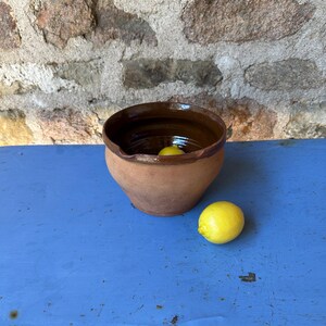 Puede incluir: Un cuenco de terracota de forma redonda con el interior esmaltado en marrón oscuro y un pequeño pico vertedor. El cuenco presenta una superficie exterior mate y sin esmaltar. Un limón amarillo brillante está colocado dentro del cuenco, y un segundo limón amarillo descansa sobre la superficie junto al cuenco.