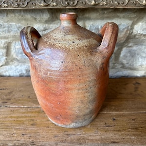 May include: A rustic, antique pottery jug with a rounded body and two arched handles. The jug has a narrow neck and a warm color palette of brown, orange, and tan. The jug is likely handmade and shows signs of age.