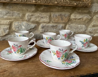 May include: Six white teacups and saucers with floral designs. The cups and saucers have a scalloped edge with a red trim. The floral pattern includes pink, red, green, and blue flowers. The set is arranged on a wooden surface.