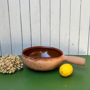 Op de afbeelding: Een ronde, ondiepe kookpan van aardewerk met één dik handvat en een kleine schenktuit. De binnenzijde is voorzien van een glanzende, donkerbruine glazuurlaag, terwijl de buitenzijde een matte, natuurlijke terracotta kleur heeft.