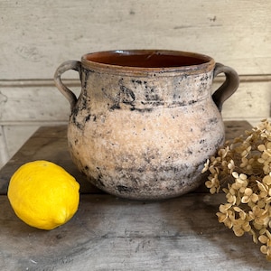 Pode incluir: Um vaso de terracota redondo e rústico com duas alças laterais, apresentando um acabamento desgastado e manchado em bege e preto. O interior é liso e castanho-avermelhado, contrastando com a superfície exterior texturizada e envelhecida.