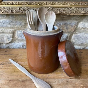 以下が含まれることがあります： 木製のスプーンとフォークが入った、蓋付きの茶色のセラミック製調理器具ホルダー。木製のスプーンがホルダーの隣の木製の表面に置かれています。 キッチン用品。