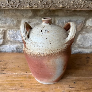 May include: A vintage stoneware jug with two handles. The jug has a speckled, off-white upper half and a reddish-brown lower half. The jug sits on a wooden surface, with a stone wall in the background.