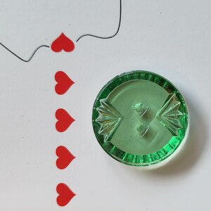 May include: A round, translucent green button featuring a central two-hole design and decorative fan-shaped embossed patterns on the sides. To the left of the button, a vertical row of five small, solid red heart shapes is arranged.