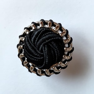 May include: A circular button featuring a central black knotted fabric dome surrounded by a silver-toned metal frame. The outer edge of the metal frame is wrapped with thin black thread, creating a detailed, textured border around the centre.