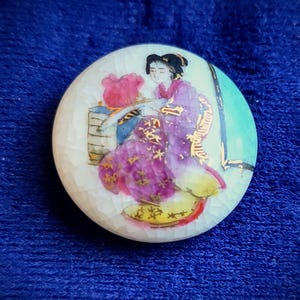 May include: A round brooch with a white crackle-finish surface featuring a painted illustration of a figure in a purple robe with gold accents. The figure is seated near a pink object and a vertical black line, set against a light green background.