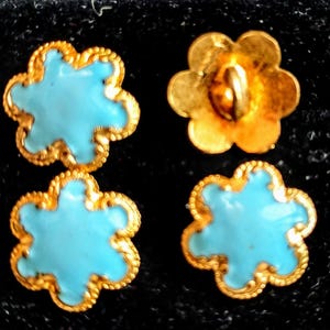 Könnte beinhalten: Vier blumenförmige Knöpfe mit einer leuchtend türkisfarbenen Emaille-Mitte und einem strukturierten goldfarbenen Metallrand. Drei Knöpfe liegen mit der Emaille-Seite nach oben, während ein Knopf umgedreht ist, um die Rückseite und den Ösensteg zu zeigen.