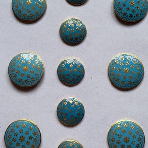 Könnte beinhalten: Eine Sammlung von neun runden Knöpfen in verschiedenen Größen mit einer leuchtend türkisblauen Emaille-Oberfläche, die mit einem wiederkehrenden Muster aus kleinen goldfarbenen Blumen und einem dünnen goldfarbenen Rand verziert ist.